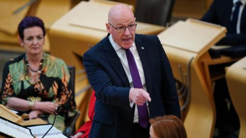 John Swinney points angrily in the chamber with Health Secretary Angela Constance sitting in the background