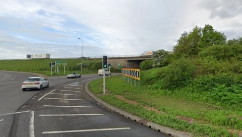 Part of the Brampton Interchange near the busy A14 and A1. There are several vehicles on the road and one on a bridge. There are traffic lights on the road and signs to exits on the road. The large roundabout has many shrubs as well as grass