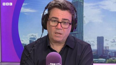 Andy Burnham sits in the Radio Manchester studio with a purple microphone in front of him, wearing glasses and over the head headphones.