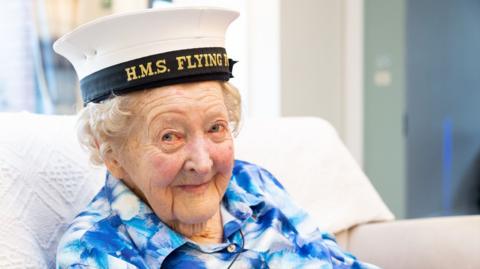 Margaret Grey sitting on a white sofa in the care home. She is wearing a blue and white patterned blouse with white buttons. On her head is the white navy cap with a black brim and yellow embroidered letters which spell out HMS Flying Fox.