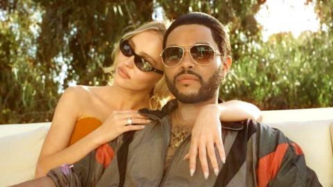 A young white woman and a slightly older black man sit on an expensive-looking sofa on a white marble veranda. They appear to be in a sunny country. Both wear sunglasses and she's sitting behind him, leaning on his shoulder. She's wearing a orange tube top and a skirt, her bare feet are curled up and resting on the edge of the couch. He's wearing a dark grey tracksuit top with black and red accents which looks retro. He's wearing trousers in a matching shade and resting one arm on the woman's knees.