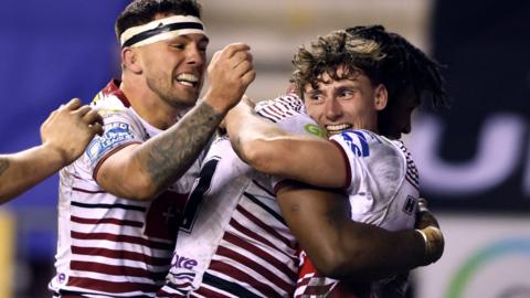 Noah Hodkinson (right, in Wigan's red-and-white kit) is hugged by Junior Nsemba after scoring a try, with two another team-mates joining the celebration