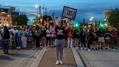 A woman in a black shirt that reads "we been not breathing" holds her fist and her hand in the air while other protesters stand in the background
