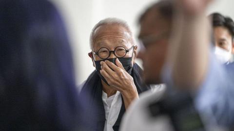 Ong Beng Seng wearing a black mask and round spectacles. He is covering his mouth with his hand.