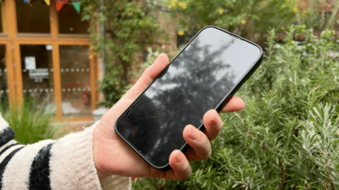 A hand holding a cracked phone, with a garden in the background.