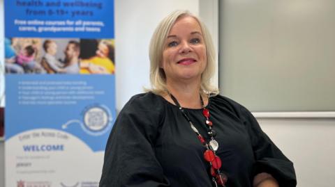 A blonde woman with a black top and large red necklace stands next to a sign which says: Free online courses for all parents, carers, grandparents and teens