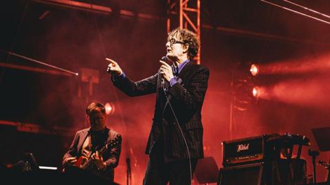 Jarvis Cocker points and sings into a microphone on stage. Next to him is a guitarist. 