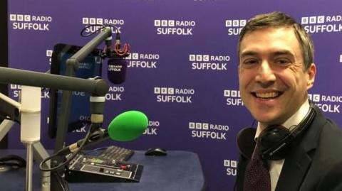 MP Adrian Ramsay is in the BBC Radio Suffolk studios sat down at the desk with a microphone ready for the BBC Radio Suffolk hot seat. He is wearing a jacket and tie and smiling at the camera. 