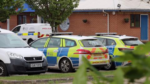 Police cars and vans parked outside Maldon and Tiptree FC's brick clubhouse.