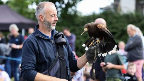 A bird handler is holding a hawk on his gloved hand. A blurred crowd is stood behind him. The hawk has food clasped in its beak.