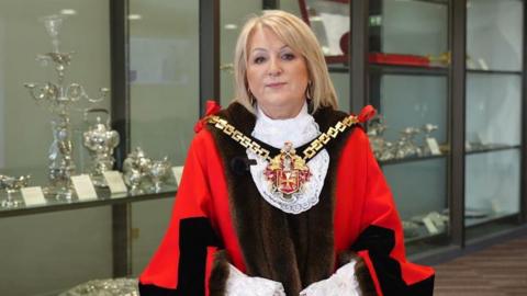 A woman with blonde hair wearing mayoral robes and chain. She is standing in front of cabinets containing the city silver.