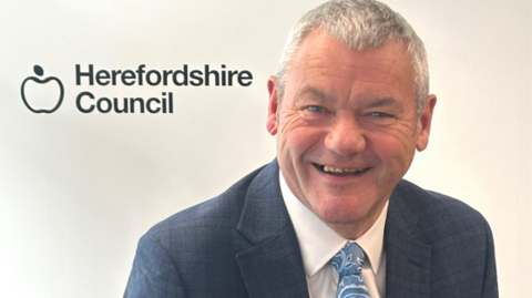 Mr Walker is wearing a navy suit, white shirt and blue patterned tie. He has short grey hair. He is smiling in the photos which also displays the council's name in the background.