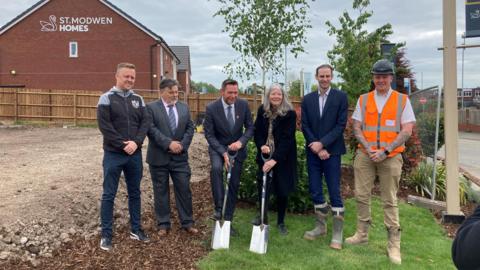 Five men and a woman stand in a line, with one of the men and the woman (both in the centre) holding spades. The man on the right is wearing a hard hat and orange hi-vis.