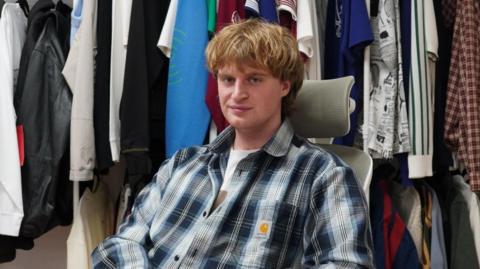 Alexei Hamblin has blond shaggy hair. He is looking at the camera and is sitting in front of a rail of clothes.