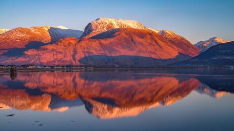 A view to Ben Nevis bathed in an orange glow. The mountain is reflected in the still waters of Loch Linnhe.