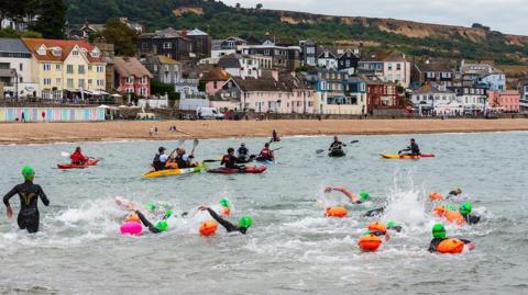 A large group of swimmers with green caps and orange floats in the sea at Lyme Regis. Nearby are about half a dozen people in kayaks and in the background is the seafront, buildings and cliffs