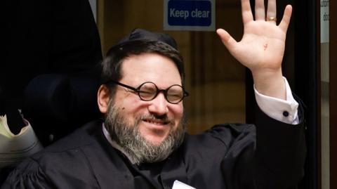 Head and shoulders of a smiling Nicholas Rossi, who is being pushed in a wheelchair. He waves and is wearing a wedding ring. He wears black circular glasses, a white shirt and what appears to be a black judges' gown.