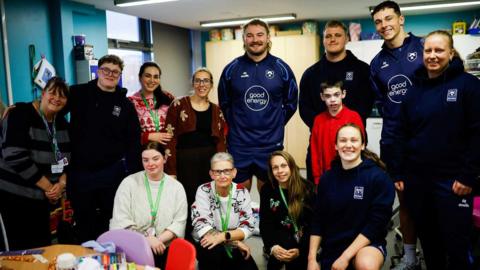 A group of about a dozen people pose for a photo with just over half of them stood behind while four women kneel in front. They are of varying ages and a mix of men and women. Some of them are from Bristol Bears.