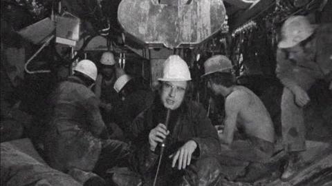 Black and white image of Bernard Falk facing the camera with a hard hat on, holding his microphone.  Behind him tunnellers work, facing away from the camera.