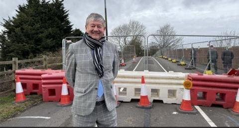 A man with short grey hair, wearing a blue and white striped scarf, and a grey checked suit (with blue tie peeping out) stands on a road in front of bollards showing the road is closed