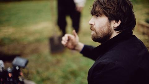 A brown haired man with a beard, wearing a dark coat in a field. He looks towards a camera operator who is slightly out of shot, giving them a thumbs up. An actor holds a shovel in the background.
