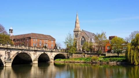 An arched stone bridge over a river with a church spire and a large brick building seen on the far side and the buildings and trees reflected in the water