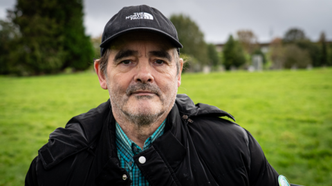a man with a short greying beard is wearing a black baseball cap and a black jacket over a green checked shirt