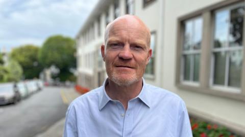 Deputy Gavin St Pier - A bald man with a ginger and grey speckled beard. He's wearing a blue buttoned shirt with the collar undone.