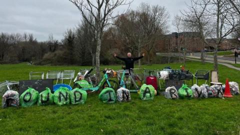 In a green park, Mark Rowlands poses with his arms out as he stands behind a row of rubbish backs with the letters 'FOUND IN A RIVER' written on them, as well as a row of shopping trolleys, e-bikes and e-scooters. 