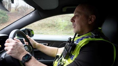 A police officer, male, wearing a luminous yellow vest with a walkie talkie attached, driving a car.