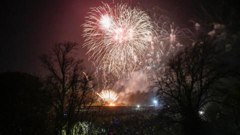 A dark sky full of fireworks with one exploding high up and a second one on the ground. They are framed by dark trees 