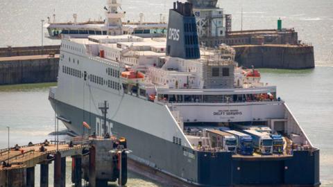 A white and navy ferry manoeuvring in a port. DFDS is painted on its funnel and the side of the boat and some lorries are visible on the deck.