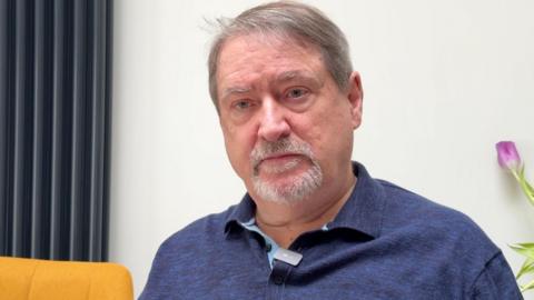 Alistair Ramsay has short grey hair and a grey goatee. He's wearing a dark blue polo neck t-shirt and sitting indoors. There's a black wall radiator to his left with the top of a yellow chair visible, and a glimpse of a purple tulip to the right.