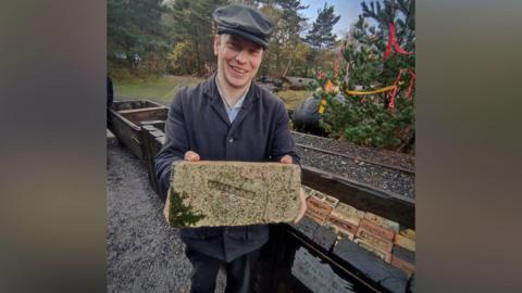Alexander Appleton is smiling as he holds a brick while standing in front of a narrow brick path at Beamish Museum. He is wearing a black buttoned-up shirt and black trousers, as well as a dark grey baker boy hat. 