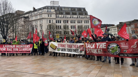 Dozens of protesters holding red unite the union flags and three large banners with messages in support of the bin strikes.