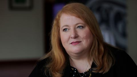 Naomi Long, a woman with long, wavy ginger hair, looks off-camera during an interview.  She is wearing a black jacket or cardigan over a multi-coloured top.