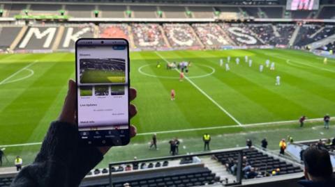 Image of someone sitting in a football stadium holding up a mobile phone, with images of a football pitch on it.  In the background can be seen a number of empty stadium seats, a football pitch, football players and officials.