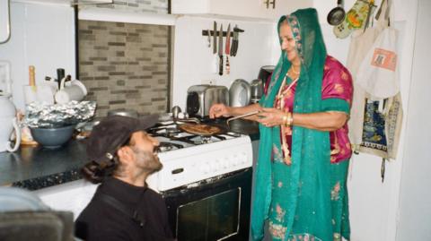 Jay Singh is sat in his grandma's kitchen, head height with the kitchen counter. He is wearing a black cap and a black turtleneck jumper. He is looking up at his grandma as she cooks on the hob while wearing green and pink traditional south Asian clothing. In the kitchen are various common kitchen item such as a kettle, toaster, oven and kitchen knives.