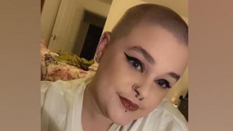 A head and shoulder picture of Tamzin Usher. She has a shaved head and blue eyes. She is wearing a white top and has piercings on her nose and lip. She has black winged eyeliner and dark lipstick. 