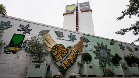 Grenfell tower covered in fabric and scaffolding, with a sign at the top that has a green heart and the phrase "Grenfell Forever in our hearts" on it. In the foreground are tribute to the victims hung and written on a nearby wall.