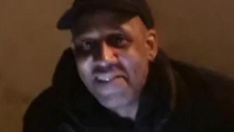A man in low lighting smiles. He is wearing a black top and a black cap.