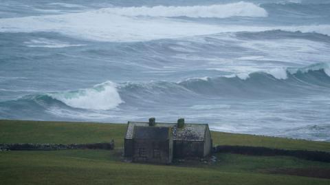 A scene of stormy seas with high waves crashing in the background. In the foreground a small house sits on green fields on top of a cliff.