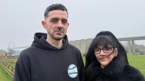 A man and a woman stand in front of a suspension bridge on a windy, foggy day. The man, on the left, is wearing a black hoodie. He has brown hair and a brown beard. The woman, on the right, is shorter and wearing a black fluffy coat. She has black hair and glasses. Only parts of the bridge are visible in the thick fog. 