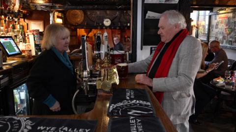 Two people in a pub. The woman behind the bar has blonde hair and a black top with turqouise top underneath. She is talking to a man with grey hair, a grey jacket and red tie