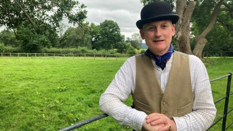 Francis Acton, wearing a bowler hat, shirt and waistcoat, and standing in a fiel