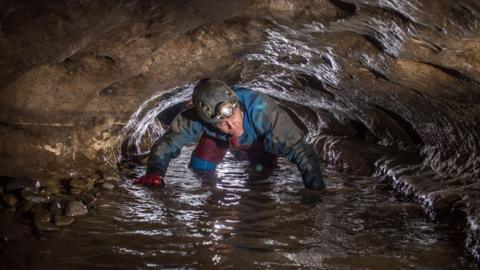 The photo shows a person wearing a helmet with a headlamp crawling through a narrow, wet cave passage, surrounded by rocky walls and water on the ground.