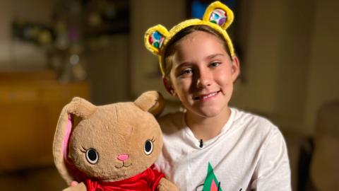 Gracie smiles at the camera wearing yellow bear ears. She is wearing a T-shirt and holding a large bunny rabbit.  