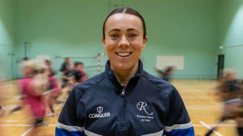 A woman smiling at the camera. She is wearing a jumper which says "Rainham School for Girls" on it. There are children running behind her.