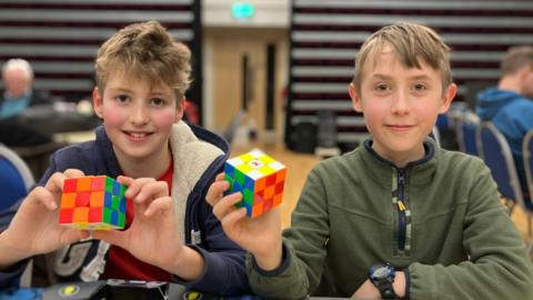 Benji and Charlie, both young boys with blond hair, at the speedcubing event. They are both holding a Rubik's cube and smiling at the camera.