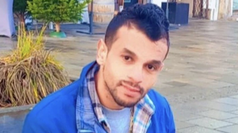 A man in a blue jacket and checked shirt sat down on a bench outside. He is holding a phone with headphones plugged in